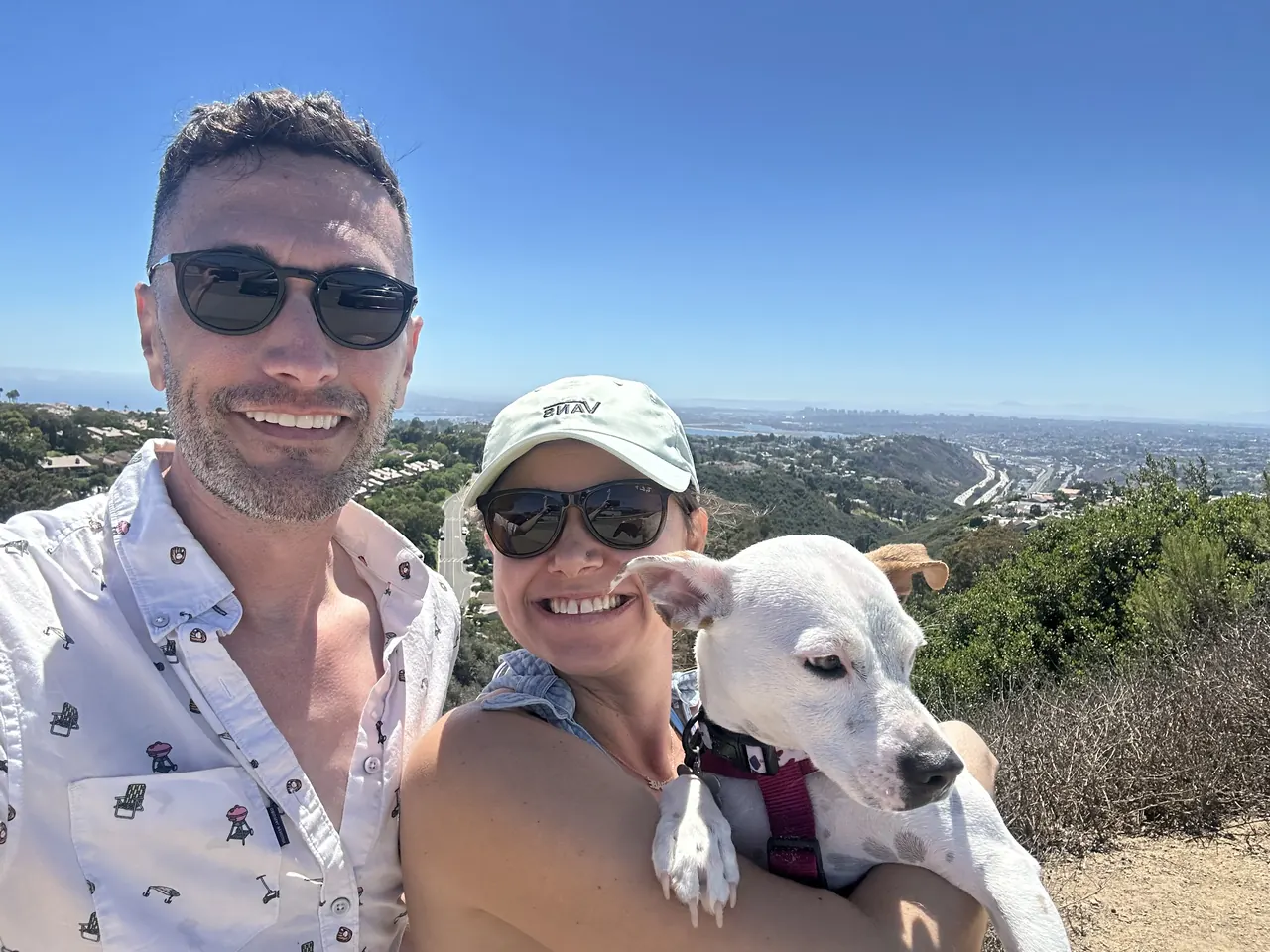 Robbie and a friend holding a white dog on a sunny hilltop overlooking the coast