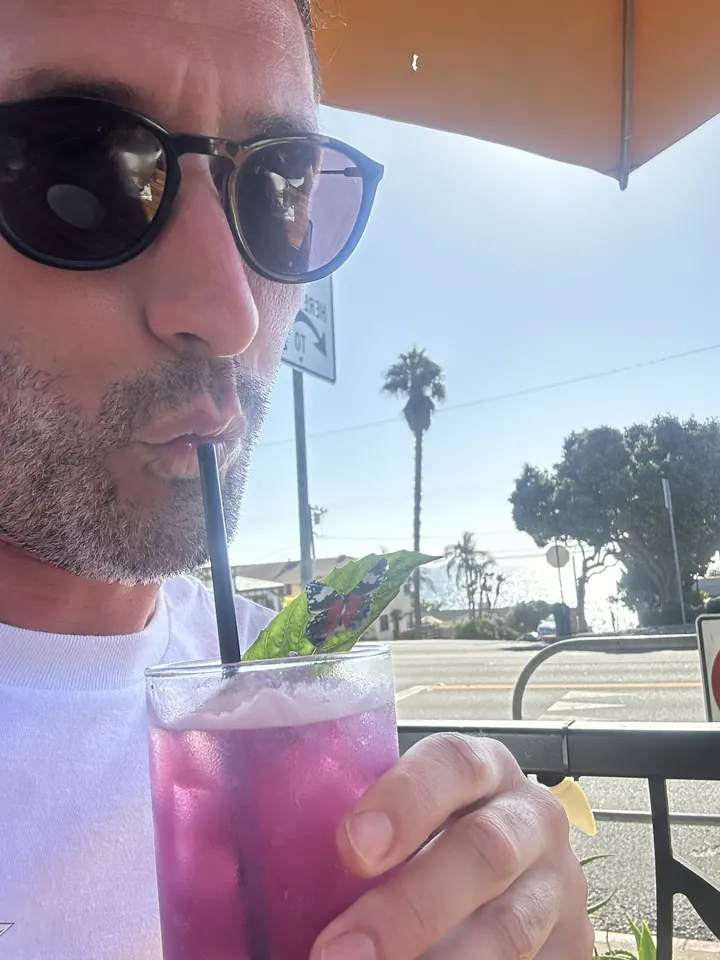 Robbie in sunglasses sipping a purple drink at a sunny outdoor table